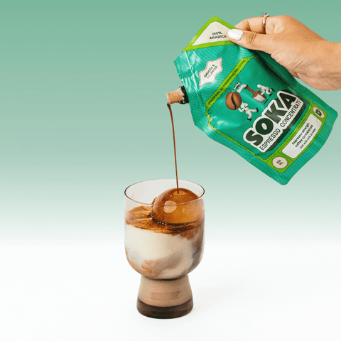 A hand with white manicured nails squeezes a green pouch of SOKA '100% ARABICA' coffee concentrate into a glass of milk with a large ice sphere, set against a light green gradient background.