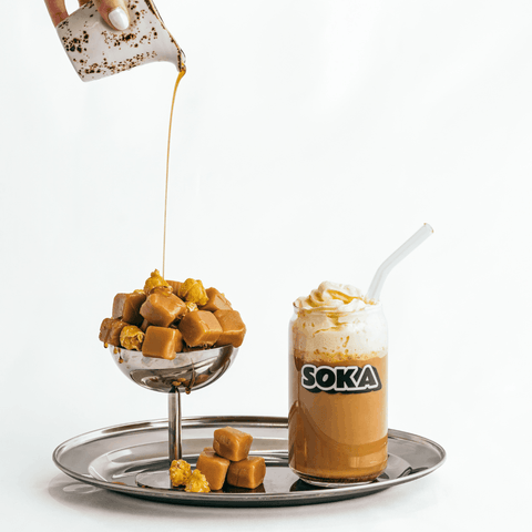 A styled beverage scene on a silver tray against a white background. One side shows caramel cubes being poured into a metal cup, and the other side shows a frosted drink topped with whipped cream and a straw in a glass labeled SOKA.