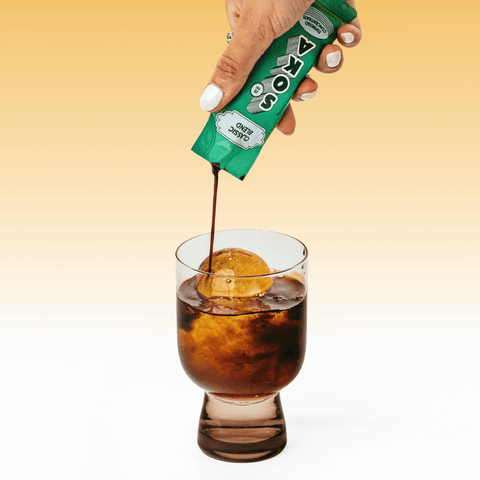 A hand pours coffee from a SOKA sachet into a glass of iced water, set against a warm yellow-to-white gradient background.