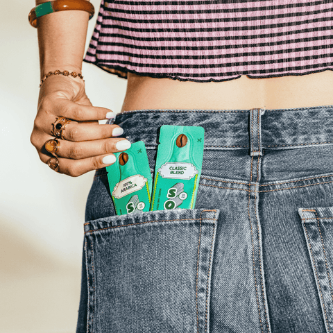 A close-up lifestyle shot of a person wearing blue denim jeans with two green coffee sachets tucked into the back pocket, labeled '100% Arabica' and 'Classic Blend'.