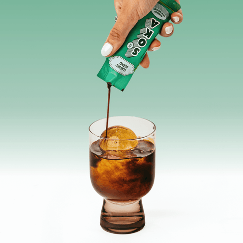 A hand with white manicured nails squeezes a green packet of SOKA espresso concentrate into a glass of water with a large ice sphere to make an americano, set against a light green gradient background.