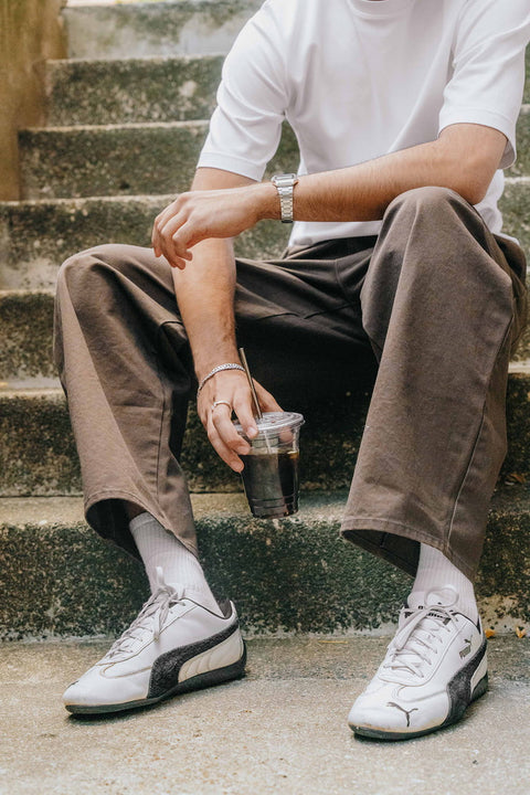 A person sitting on outdoor concrete steps wearing a white t-shirt, brown pants, white socks, and white Puma sneakers, holding a clear plastic cup of iced coffee.