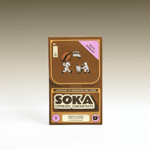 A box of SOKA Espresso Concentrate coffee sachets, featuring a brown and tan design with cartoon illustrations of coffee beans and characters, labeled with a purple 'NUTTY HAZELNUT' sticker, set against a neutral gradient background.
