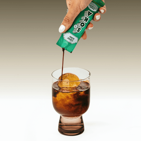 A hand with white manicured nails pours a dark brown liquid from a green SOKA sachet into a glass of iced water to make an americano coffee, set against a clean grey-to-white gradient background.