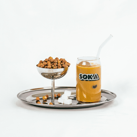 A glass of iced coffee with a clear glass straw and a visible SOKA logo, served on a silver tray alongside a small bowl of roasted hazelnuts and white sugar cubes against a clean white background.