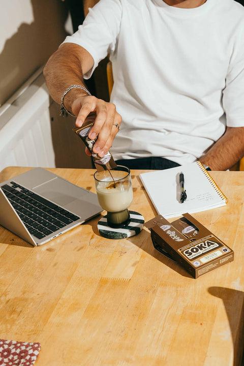 A person in a white t-shirt pours coffee from a portable sachet into a glass of milk-based drink on a wooden table, with a laptop, notebook, and a box of SOKA espresso concentrate nearby.