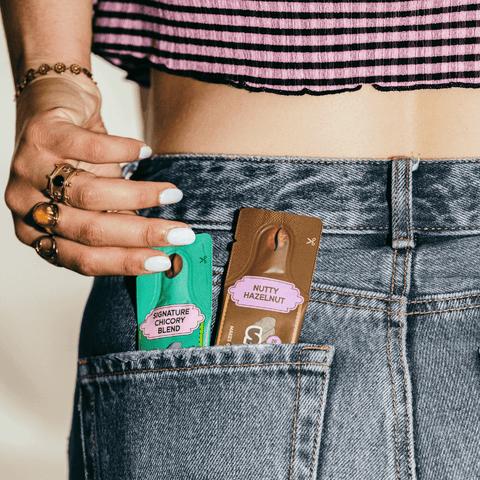 A close-up lifestyle shot of a person's hand and midsection, wearing blue denim jeans with two coffee sachets tucked into the back pocket. The products are packaged in green and brown sleeves labeled 'Signature Chicory Blend' and 'Nutty Hazelnut'.