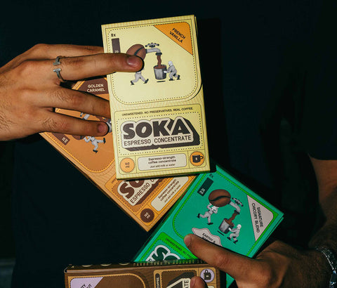 A person in a black t-shirt holds four SOKA branded boxes in their hands. The boxes are stacked and fanned out, showing different coffee flavours: French Vanilla, Golden Caramel, Signature Chicory Blend,  and Nutty Hazelnut.