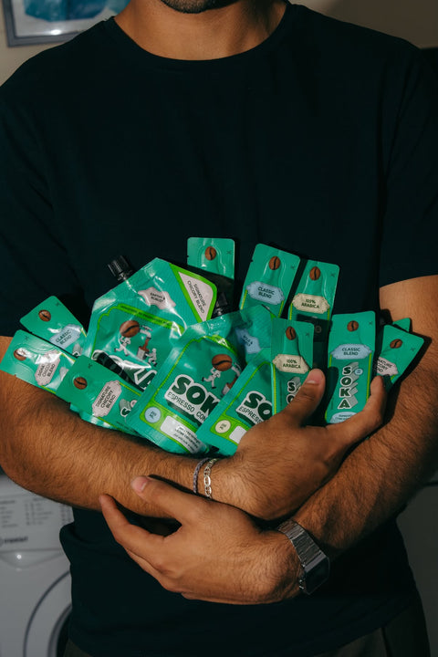 A person in a black t-shirt holds a cluster of green coffee sachets. The individual sachets feature the brand name 'SOKA' and depict coffee bean graphics.