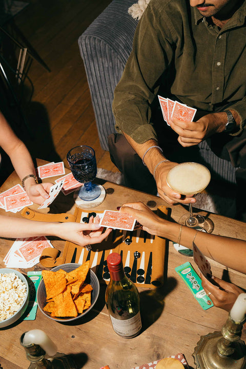 Overhead view of a group of people playing cards around a wooden table with snacks, drinks, SOKA espresso concentrate sachets, and a game board in a dimly lit indoor setting.