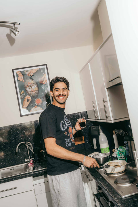A smiling young man with a moustache stands in a kitchen holding a mug of coffee. He is wearing a black graphic t-shirt and light grey sweatpants. On the kitchen countertop stands a green SOKA espresso concentrate pouch.
