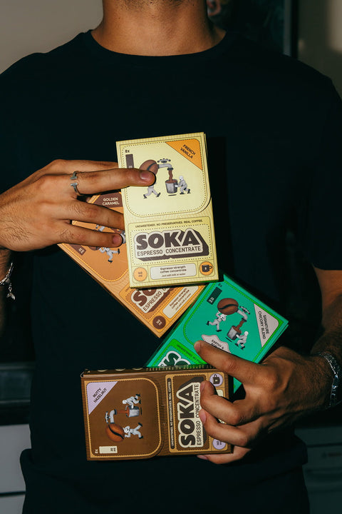 A person in a black shirt holds four SOKA espresso concentrate boxes in different colours (yellow, orange, green, and brown).