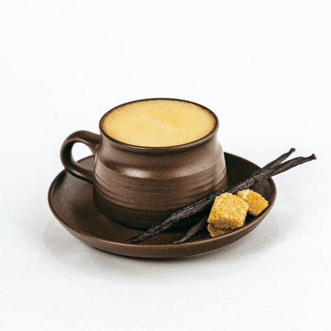 A dark brown ceramic coffee cup filled with a frothy latte sits on a matching saucer, styled with two brown sugar cubes and vanilla bean sticks on a clean white background.
