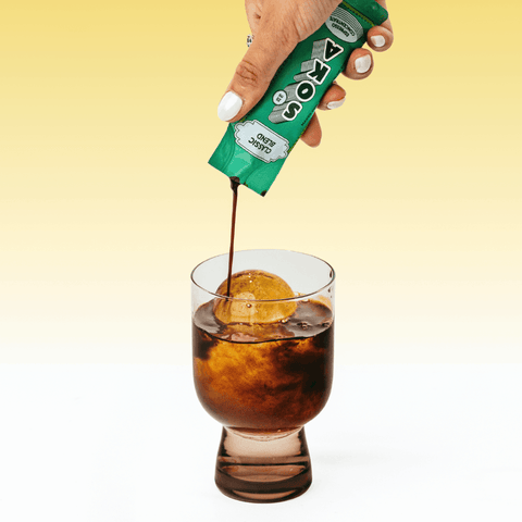 A hand with white manicured nails pours coffee from a SOKA sachet into a clear glass containing a large ice cube, set against a yellow-to-white gradient background.